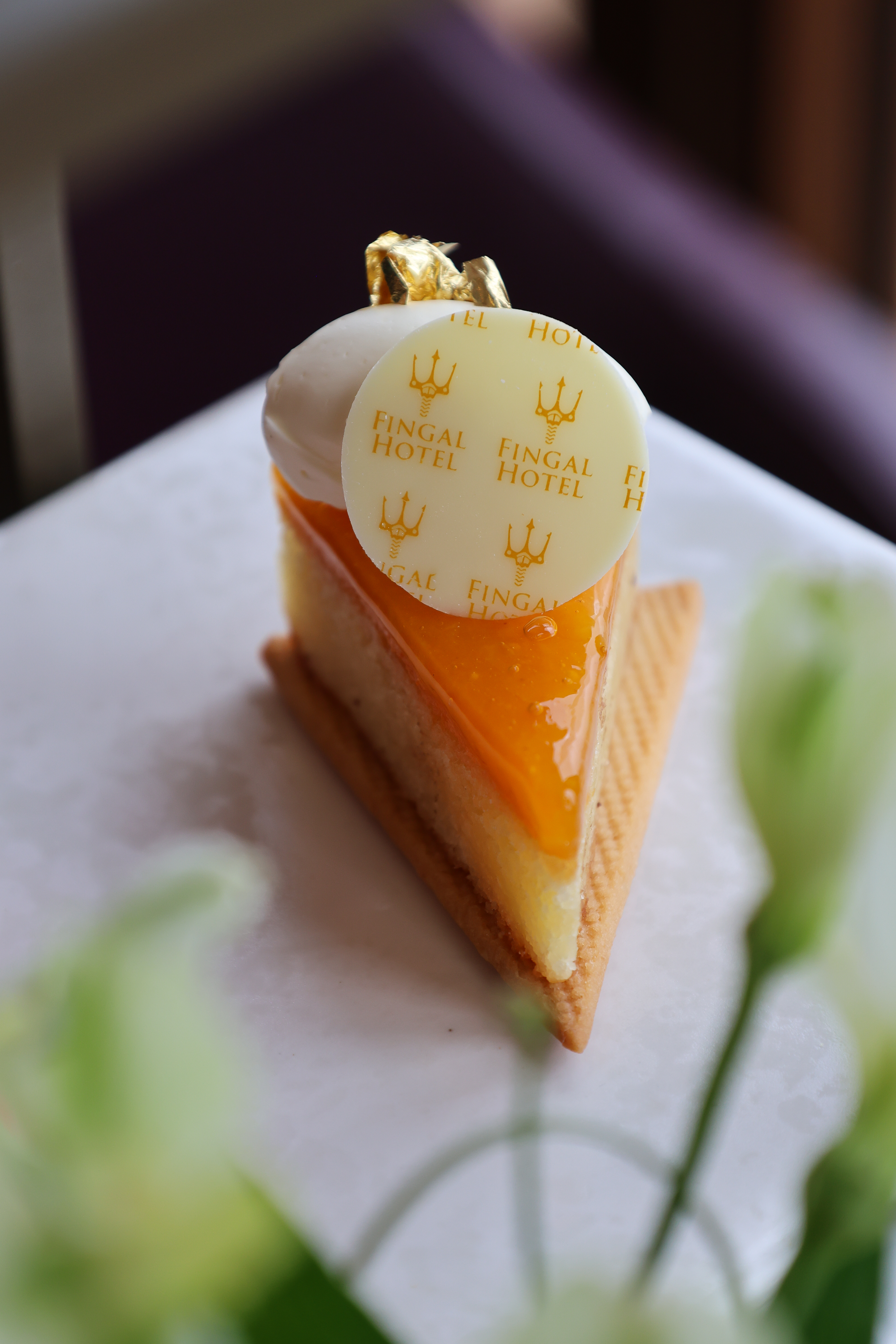 Slice of orange‑glazed patisserie on a biscuit base, topped with a Fingal Hotel chocolate disc and gold leaf.