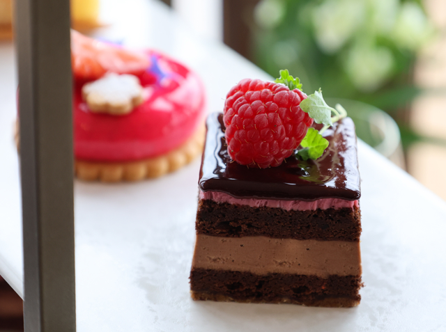Close‑up of a chocolate mousse cake topped with a raspberry and mint, with a pink glazed dessert blurred behind.