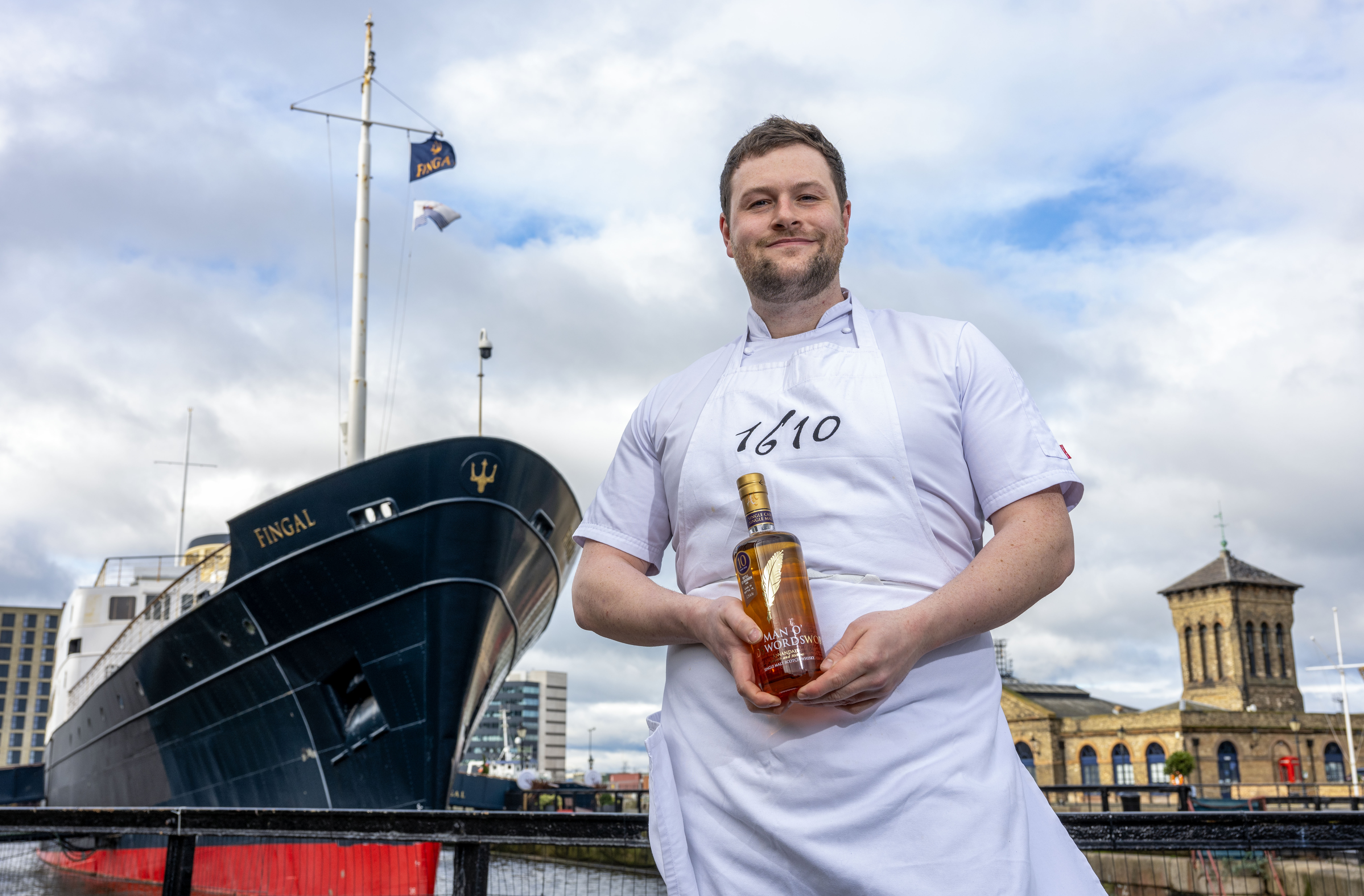 Chef Fraser stands holding a bottle of whisky in front of ship hotel Fingal