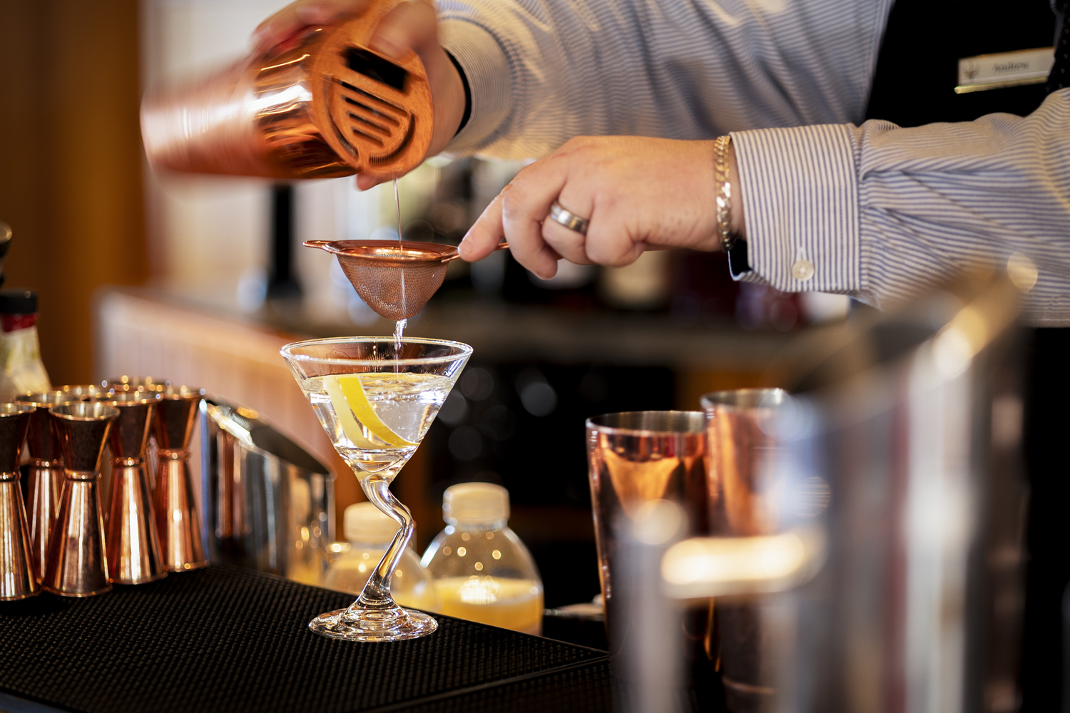 Bartender pouring a cocktail through a strainer into a martini glass, surrounded by copper bar tools.