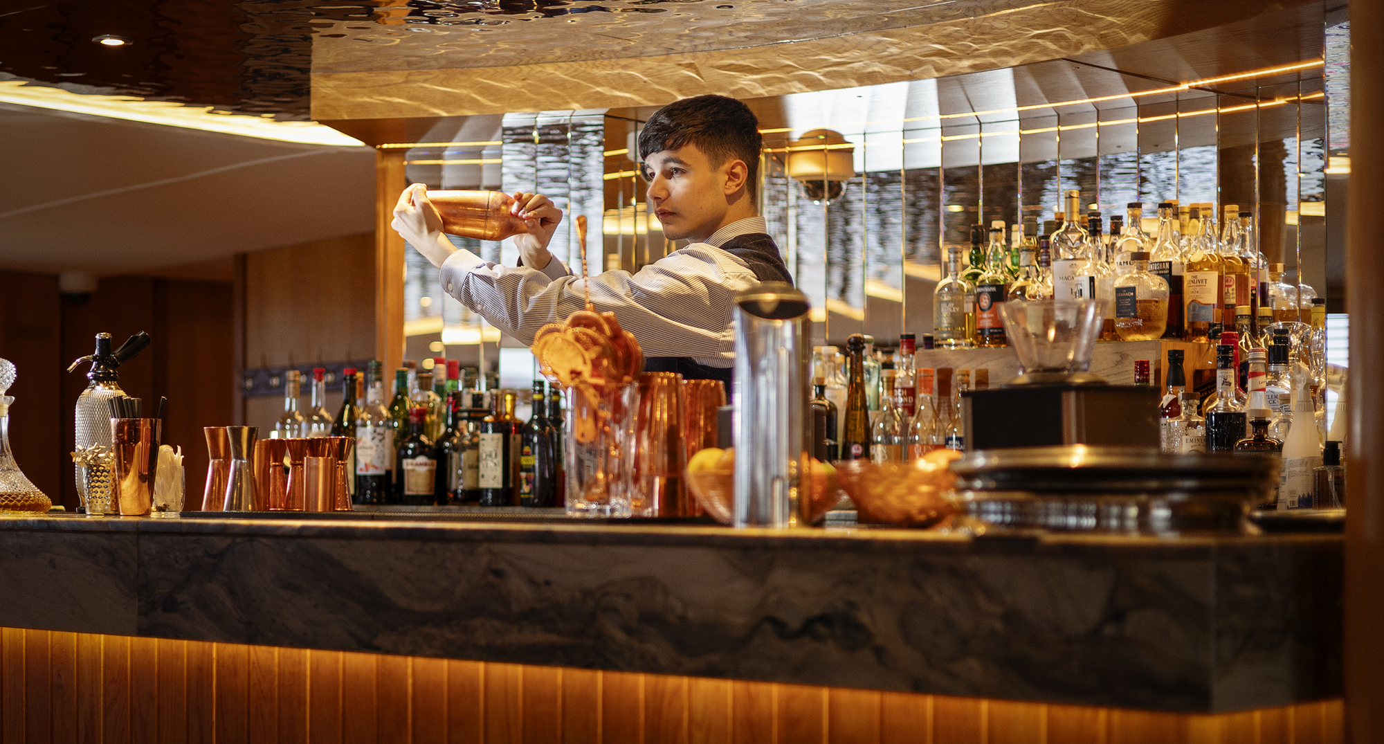 Bartender shaking a cocktail behind a well‑lit bar stocked with bottles and copper barware.
