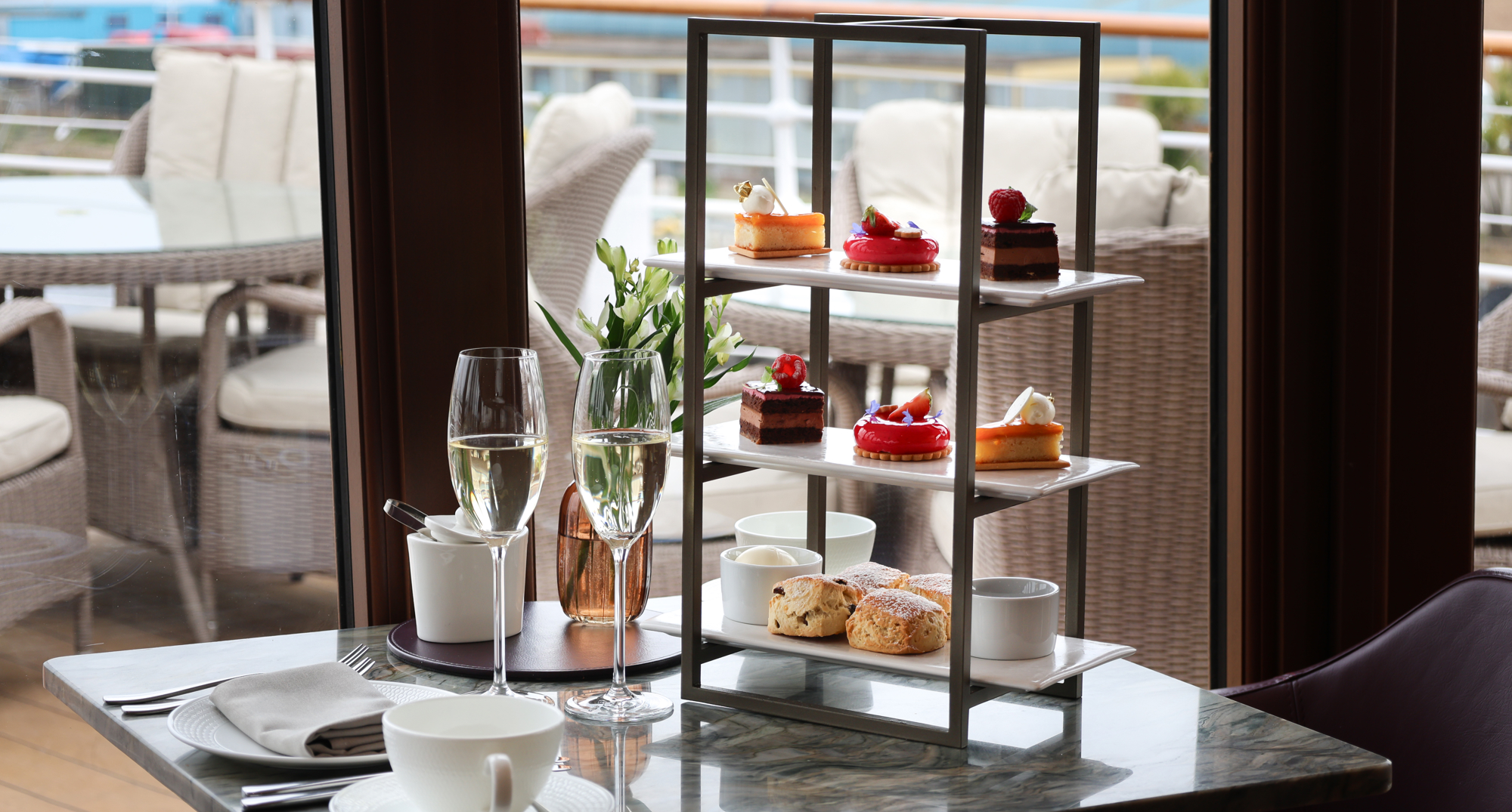 Three‑tier afternoon‑tea stand with pastries and scones on a marble table overlooking the waterfront.