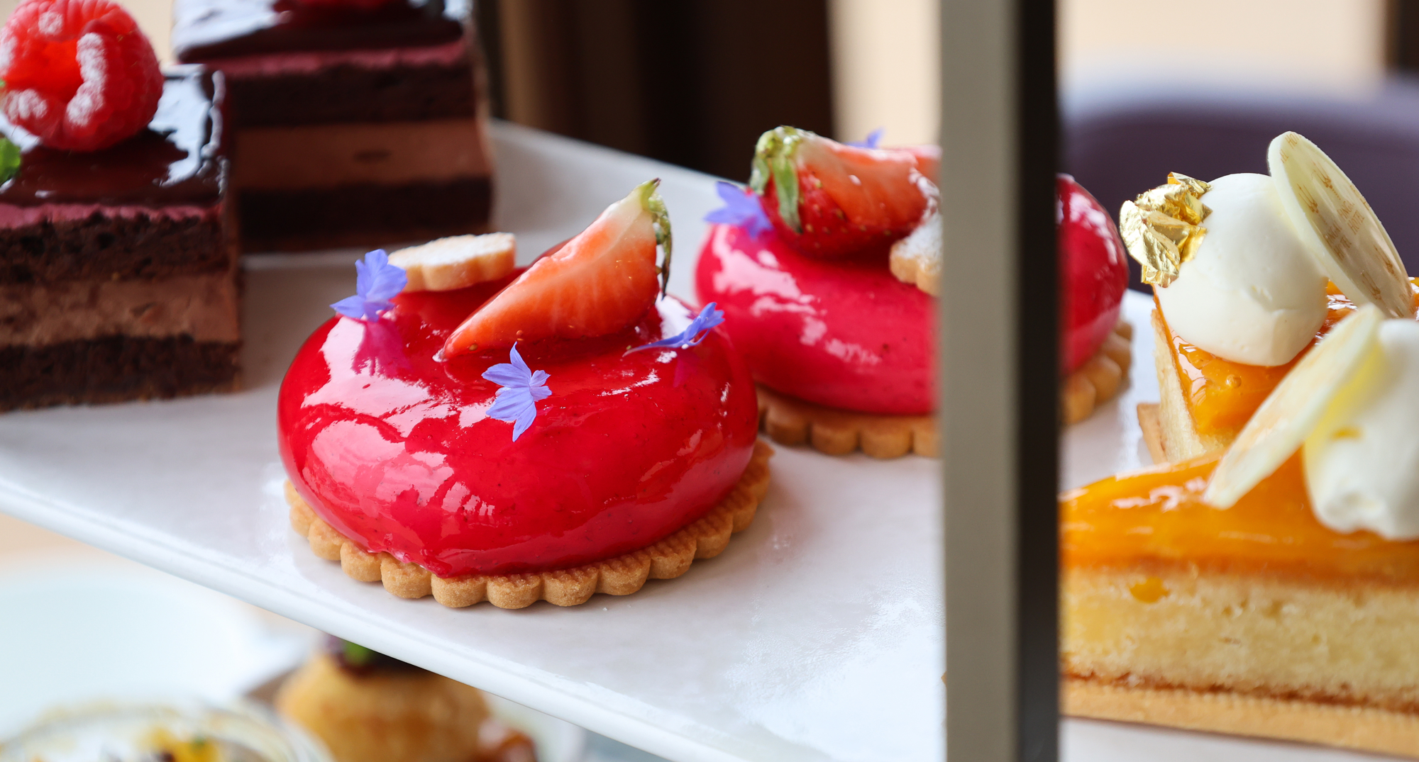 Assortment of colourful patisserie desserts on a tiered stand, including red domes, chocolate slices, and citrus‑glazed cakes.