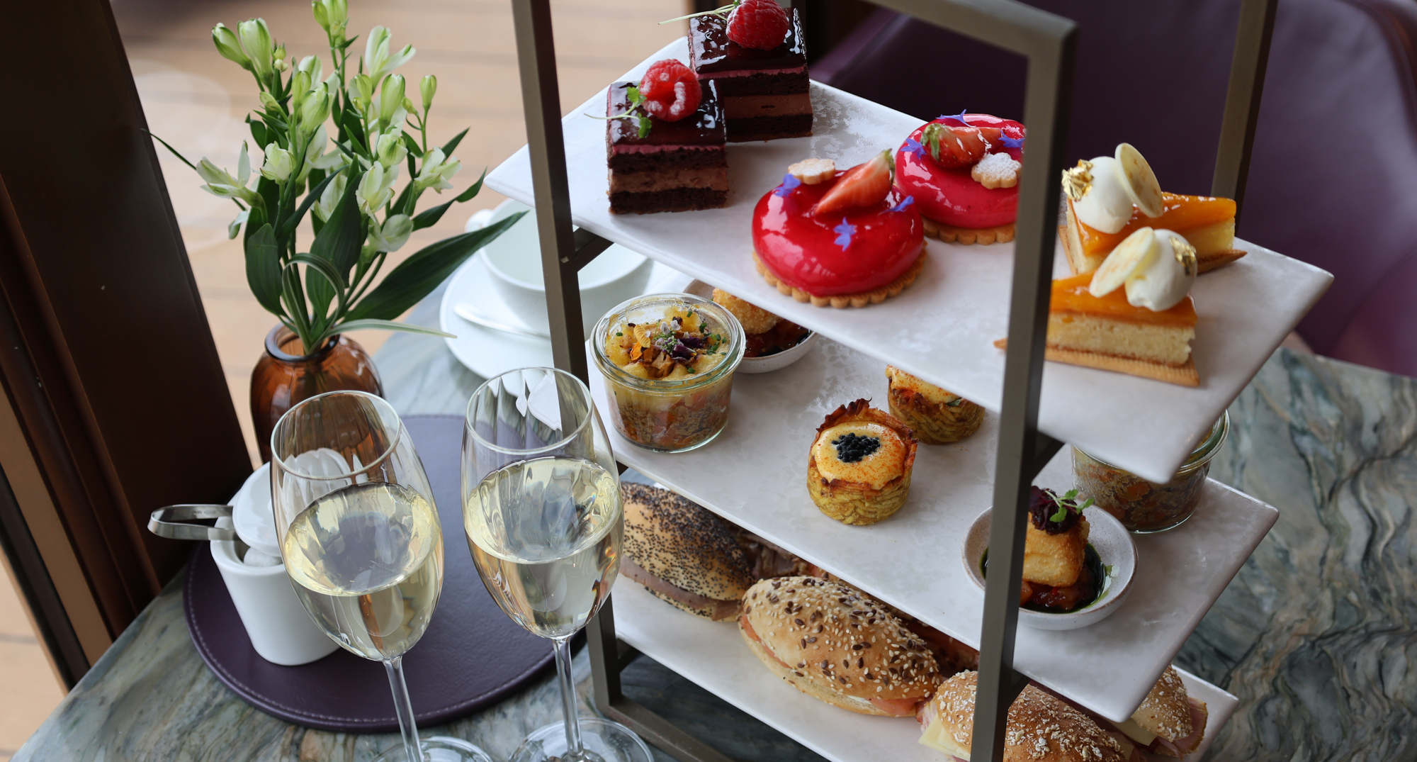 Three‑tier stand with colourful pastries, layered parfaits, and mini sandwiches, served with champagne in a restaurant.