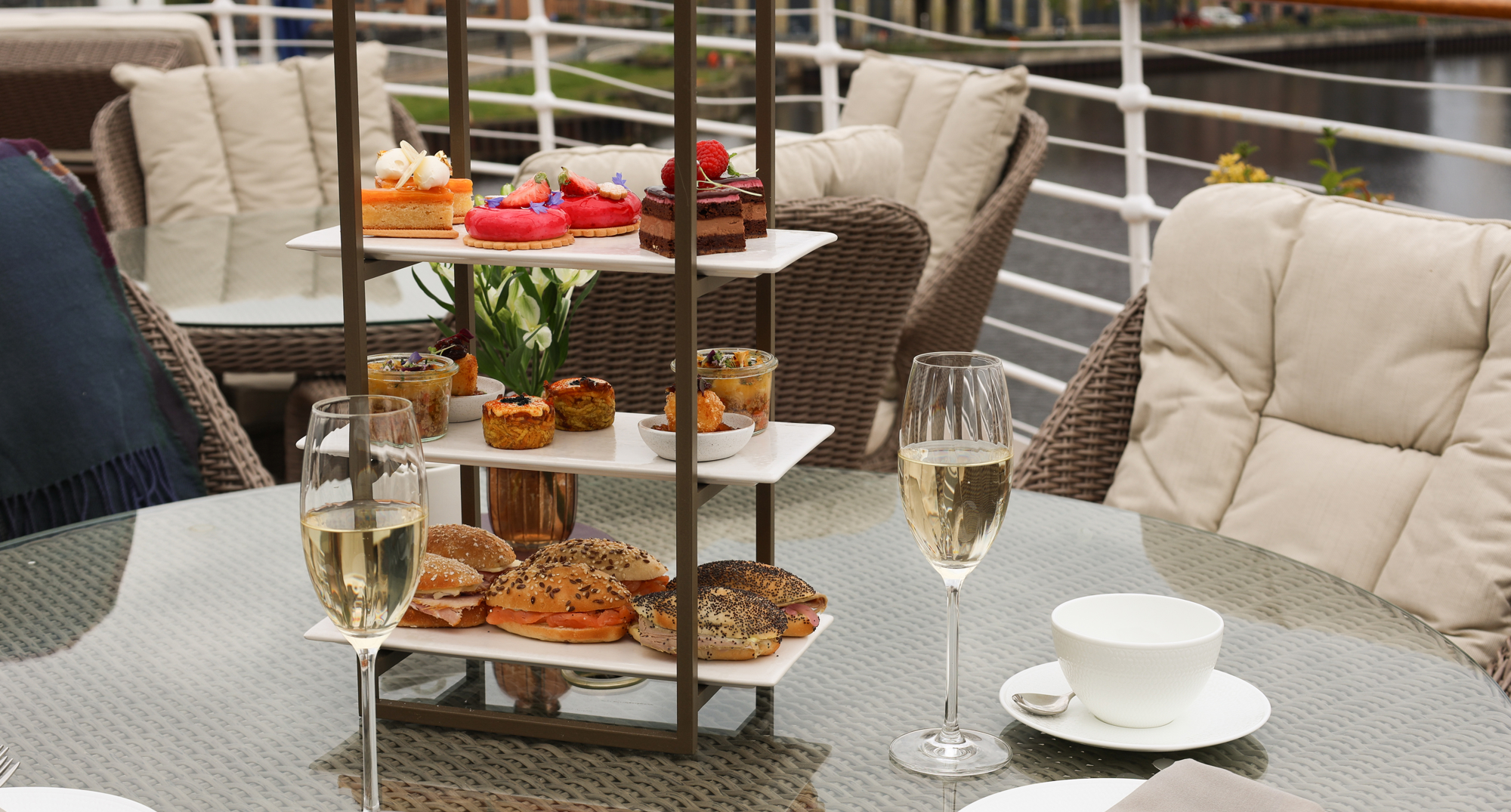 Outdoor table set with a three‑tier afternoon‑tea stand, champagne glasses, and waterfront views.